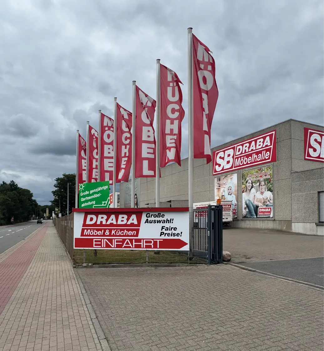 Einfahrt zur SB DRABA Möbelhalle mit roten Fahnen, Schild und Gehweg an der Straße